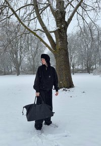 Svart hettegenser og bukser, holder en stor svart bag. Snødekt landskap med et tre i bakgrunnen. Lett snøfall synlig.