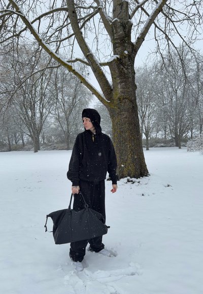 Sudadera con capucha negra y pantalones, sosteniendo una gran bolsa negra. Paisaje nevado con un árbol al fondo. Nieve ligera visible.