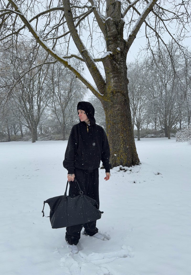 Felpa nera con cappuccio e pantaloni, che tiene una grande borsa nera. Paesaggio innevato con un albero sullo sfondo. Leggera nevicata visibile.