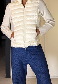 Άτομο που φοράει κρεμ μπουφάν με φερμουάρ, με πλεκτά μανίκια και μπλε παντελόνι με σχέδιο paisley, με τα χέρια στις τσέπες του μπουφάν.