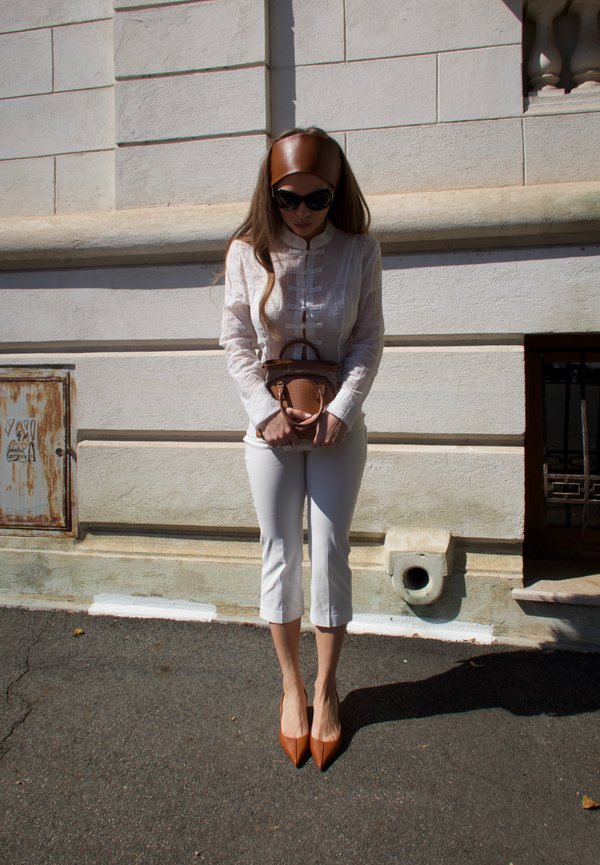 Femme portant une tenue blanche et des accessoires marron, debout contre un mur de pierre claire, tenant un petit sac marron.