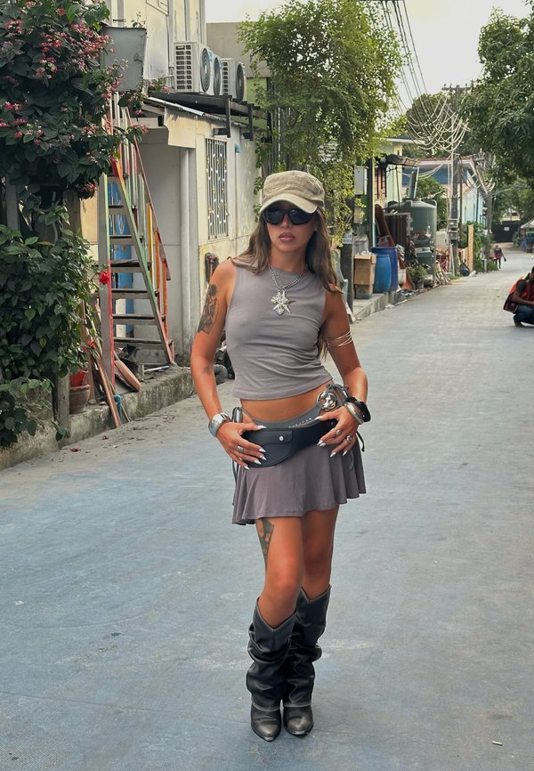 Γυναίκα που φορά γκρι αμάνικη μπλούζα και φούστα, μαύρες μπότες, γυαλιά ηλίου και καπέλο, στεκείται σε ήσυχο δρόμο με φυτά και κτίρια κοντά.