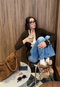 Žena s naočalama, u smeđem džemperu i trapericama sjedi na stolici držeći zeleni napitak, pored nje na stolu nalaze se smeđa torba, novčanik, ruž za usne i sunčane naočale.