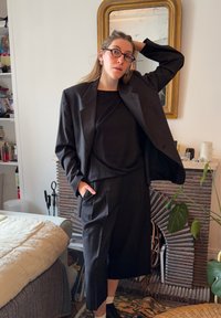 Blazer noir sur mesure et pantalon large assorti, présentant une texture douce et un design minimaliste. Stylé avec un haut noir et des chaussures noires.