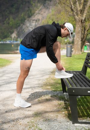 Zapatillas deportivas blancas con parte superior texturizada, combinadas con pantalones cortos azules y una chaqueta ligera negra, sobre un banco del parque junto a un sendero.
