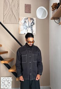 Μαύρο oversized πουκάμισο με λευκές αντίθετες ραφές, διαθέτοντας δύο μπροστινές τσέπες και σχέδιο με κουμπιά, φορεμένο πάνω από χαλαρούς μαύρους παντελόνες.