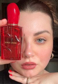 Giorgio Armanin punainen hajuvesipullo veistoksellisella muotoilulla. Kirkasta lasia, kiiltävän punainen korkki ja kultainen logoyksityiskohta. Kirkkaan punaiset kynnet etualalla.