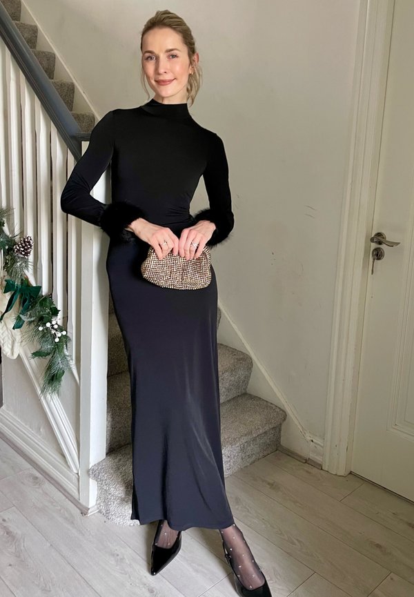 Vestido negro de manga larga con puños de piel sintética, con un diseño ajustado. Lleva un bolso de mano texturizado; viste medias negras transparentes y tacones de punta.