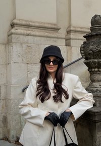 Blazer blanc sur mesure, chapeau seau côtelé noir, lunettes de soleil noires et gants en cuir. Cheveux longs et ondulés, tenant un sac à main noir.