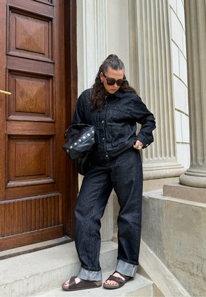 Chaqueta y pantalones de mezclilla negra, tela texturada, corte relajado, combinados con sandalias marrones. Los accesorios incluyen un bolso negro y gafas de sol.