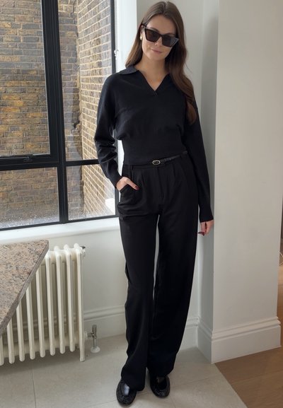 Mujer con gafas de sol negras, camiseta negra de manga larga, pantalones negros de cintura alta con cinturón, de pie en el interior cerca de una ventana y un radiador.
