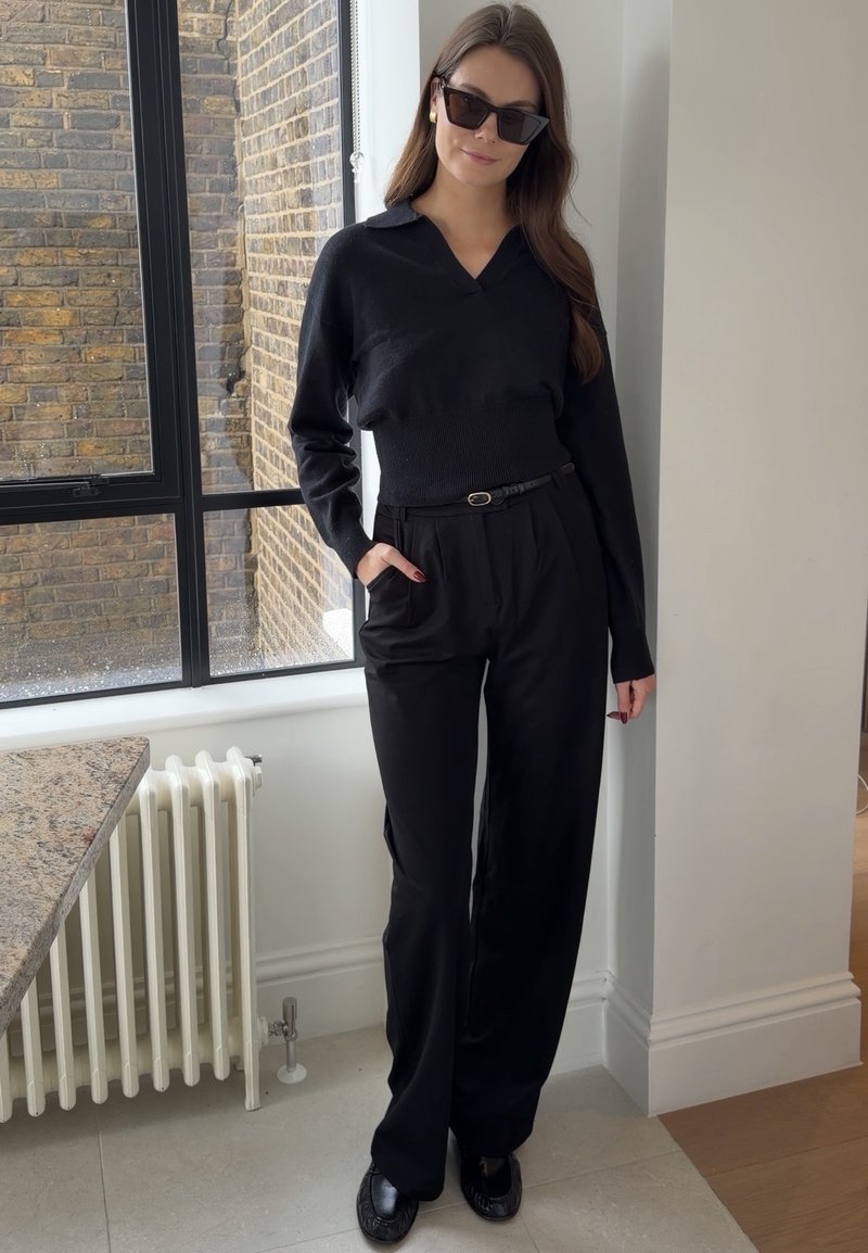 Femme portant des lunettes de soleil noires, un haut noir à manches longues, un pantalon noir taille haute avec ceinture, debout à l'intérieur près d'une fenêtre et d'un radiateur.