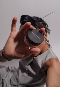 Un pot de BareMinerals Original tenu dans une main. Le couvercle noir porte un texte blanc. Le pot a une base transparente, reflétant la lumière environnante.