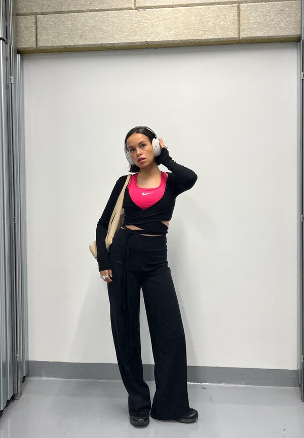 Young woman wearing white headphones, a pink Nike top, black wrap sweater and trousers, standing against a white wall with one hand on headphones.