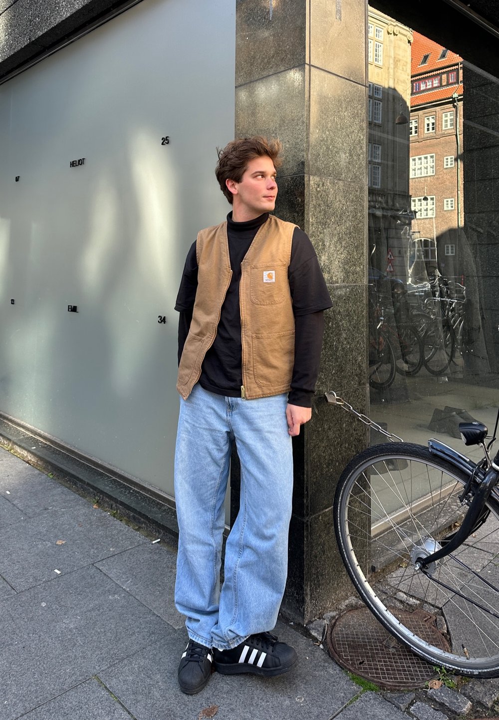 Un gilet senza maniche marrone sopra una maglietta nera, abbinato a jeans blu larghi e scarpe nere con strisce bianche, in piedi accanto a una bicicletta.