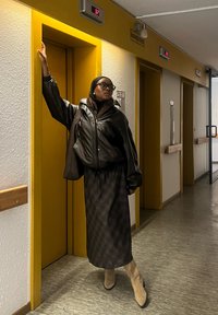 Osoba v černé kožené oversized bundě a dlouhé kárované sukni stojí u žlutého výtahu, celý outfit doplňují vysoké béžové boty.