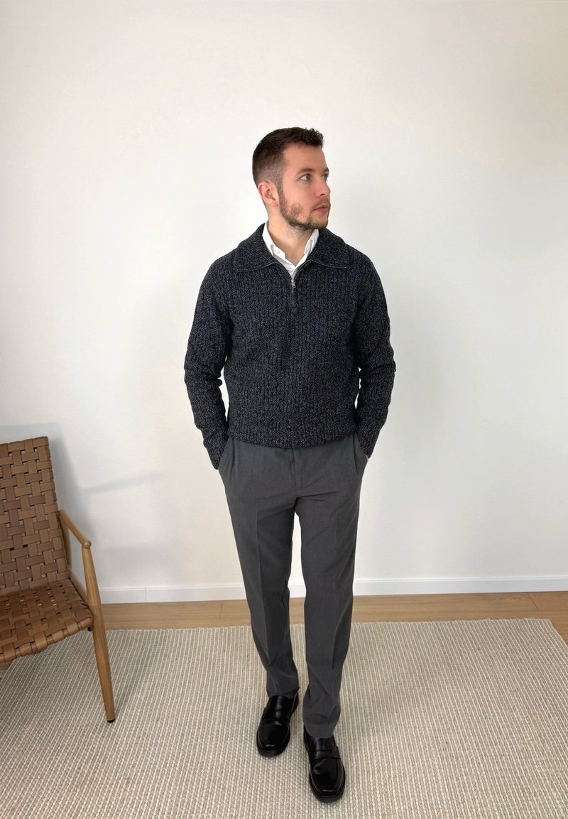 Man in dark gray sweater, white collared shirt, gray trousers, and black shoes standing on a rug next to a brown woven chair.