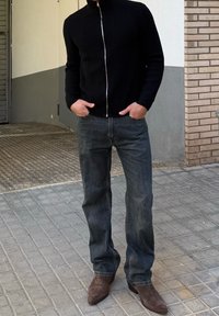 Homme portant un pull à fermeture éclair noir, un jean foncé et des bottes brunes, debout sur un trottoir carrelé gris près du coin d'un bâtiment, les mains dans les poches.