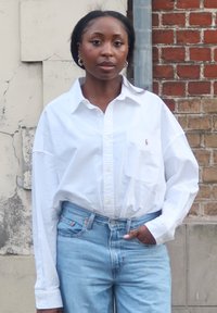 Biała koszula na guziki z długimi rękawami i kieszenią na piersi, zestawiona z jasno niebieskimi dżinsami. Model stoi na tle ściany z cegieł i o fakturze.