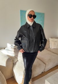 Veste courte en simili cuir noir avec col, deux poches avant et poignets élastiqués. Portée par-dessus une jupe noire avec des accents en dentelle.