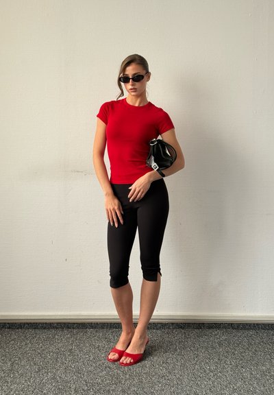 Mujer con camiseta roja, pantalones capri negros, tacones rojos y gafas de sol negras sosteniendo un bolso pequeño negro contra una pared lisa.