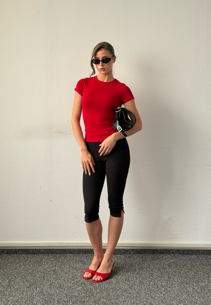 Femme portant un haut rouge, un pantalon capri noir, des talons rouges et des lunettes de soleil noires tenant un petit sac à main noir contre un mur uni.