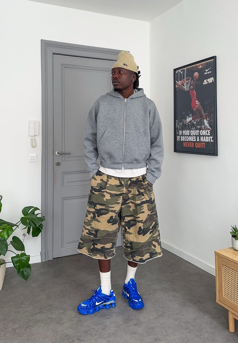 Uomo che indossa un berretto beige, felpa grigia con cappuccio, pantaloncini mimetici e sneaker blu vivace, in piedi all'interno vicino a una porta grigia e a un poster motivazionale di basket.