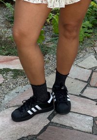 Adidași negri cu dungi albe, cu tălpi din cauciuc texturat, asortați cu șosete negre, strălucitoare, pe un fundal de alee din piatră.