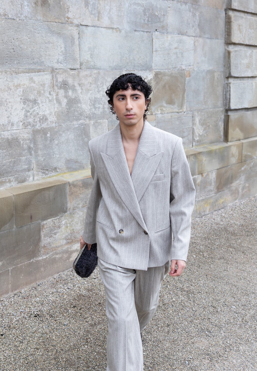 Joven caminando junto a un muro de piedra, vestido con un traje gris claro a rayas y sujetando un pequeño bolso negro texturizado.