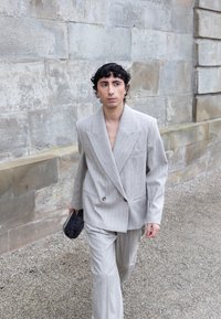 Joven caminando junto a un muro de piedra, vestido con un traje gris claro a rayas y sujetando un pequeño bolso negro texturizado.