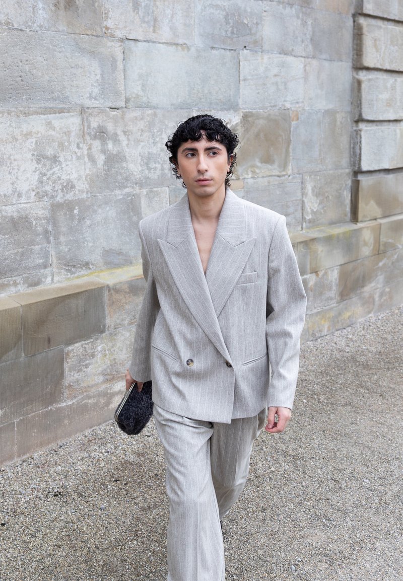 Jeune personne marchant le long d'un mur de pierre, portant un costume gris clair à fines rayures et tenant une petite pochette noire texturée.