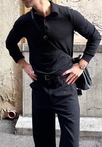 Homme portant une chemise noire à manches longues et un pantalon, debout mains sur les hanches, avec une montre-bracelet et un sac porté en bandoulière, devant un mur de pierre avec des graffitis.