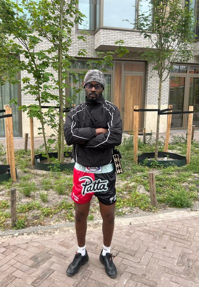 Look atlético: chaqueta negra, pantalones cortos estampados en rojo y negro con el logo de "Patta", zapatos negros, posando al aire libre junto a vegetación y un edificio moderno.