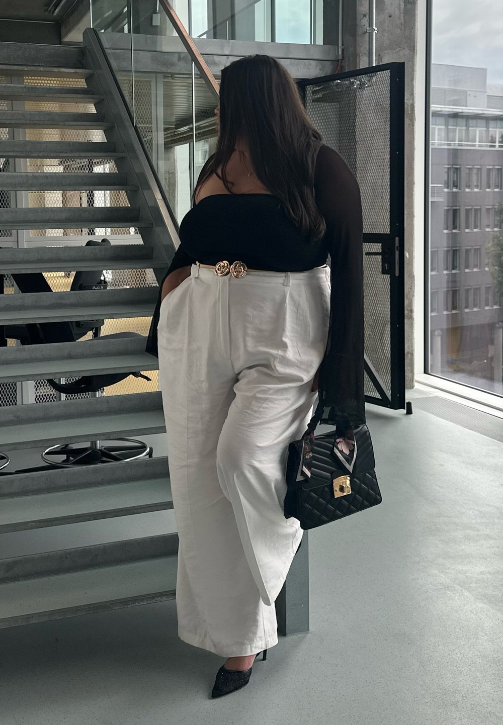 Femme debout près d'un escalier en métal, portant un haut noir à épaules dénudées, un pantalon blanc à jambes larges, tenant un sac à main noir matelassé avec un foulard floral.