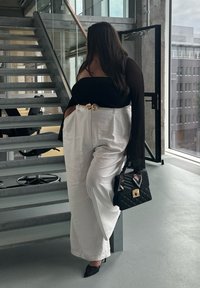 Femme debout près d'un escalier en métal, portant un haut noir à épaules dénudées, un pantalon blanc à jambes larges, tenant un sac à main noir matelassé avec un foulard floral.