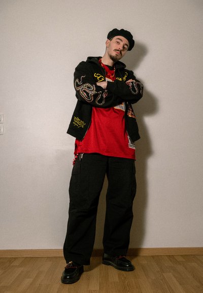 Sudadera negra de gran tamaño con diseños en amarillo y rojo, camiseta roja, pantalones cargo negros y zapatos negros. Los accesorios incluyen un collar de cuentas.