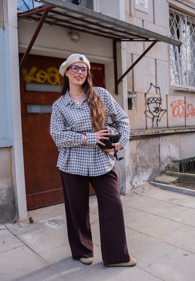 Mujer con camisa de cuadros, boina beige y gafas, de pie en una acera de la ciudad sosteniendo una bolsa negra, con grafiti en el edificio detrás.