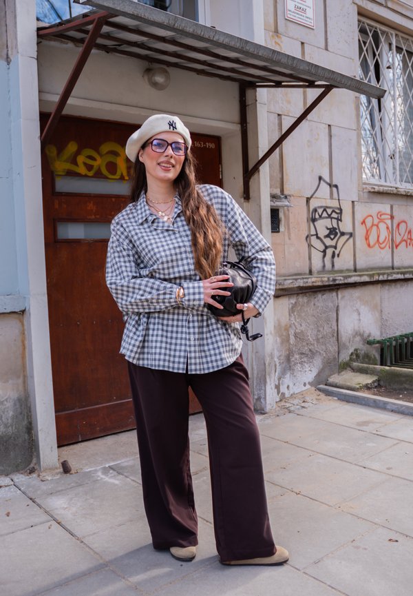 Žena u karo košulji, bež beretki i naočalama stoji na gradskom pločniku držeći crnu torbicu, a iza nje na zgradi je grafit.