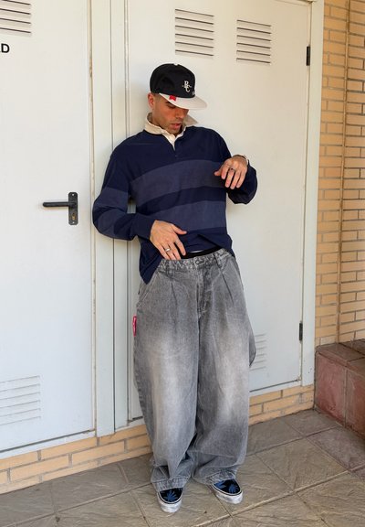 Joven con vaqueros grises de gran tamaño, suéter azul marino con rayas, gorra de béisbol negra y zapatillas negras con detalles azules, apoyado en una puerta blanca.