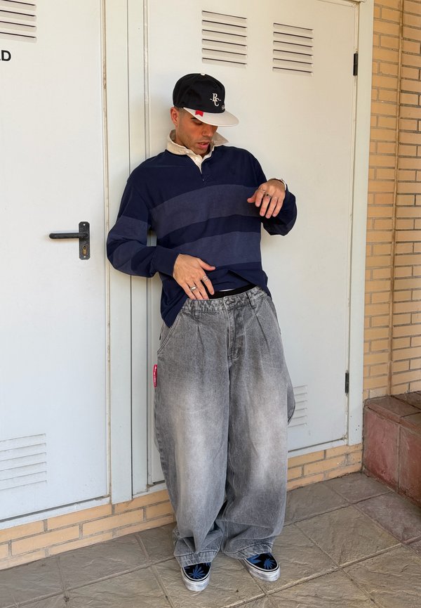Nuori mies, jolla on ylisuuria harmaita farkkuja, laivastonsininen raidallinen neulepaita, musta baseball-lippis ja mustat tennarit sinisin kuvioin, nojautuu valkoista ovea vasten.