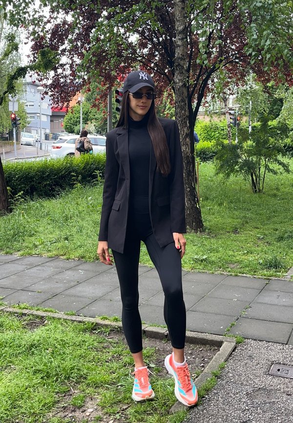 Mujer con blazer negro, leggings, gorra y gafas de sol está de pie sobre el césped cerca de un árbol junto a una acera en una zona urbana verde.