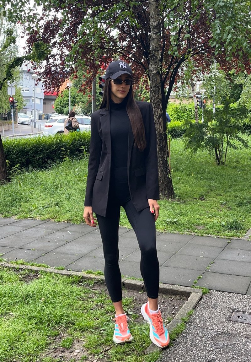 Frau mit schwarzem Blazer, Leggings, Cap und Sonnenbrille steht auf Gras nahe einem Baum neben einem Gehweg in einer grünen Stadtzone.