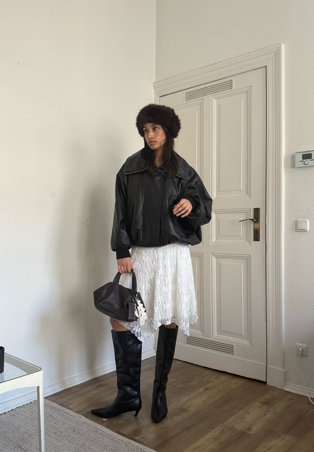 Casaco amplo de couro preto, vestido branco de renda, botas pretas até ao joelho, mala castanha, chapéu de pele; texturas suaves e cores contrastantes.