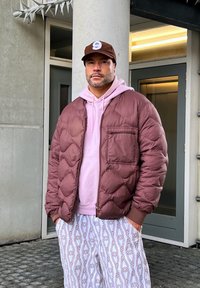Smeđa puf jakna s kvadratnim uzorkom, ružičasta dukserica ispod i bijele hlače s uzorkom. Outfit uključuje smeđu kapu s logom.