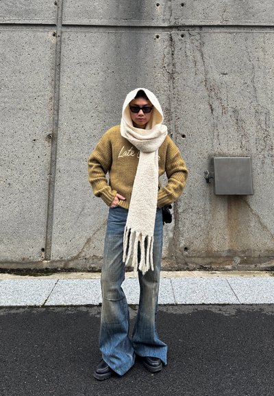Cálido suéter mostaza con el texto "Tarde", bufanda gruesa color blanco roto, jeans de pierna ancha y botas negras, frente a una pared de concreto texturizado.