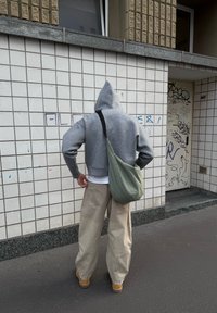 Grauer Kapuzenpullover mit Kapuze, beiger weiter Hose und einer hellgrünen Umhängetasche. Steht an einer gefliestem Wand auf dem Gehweg.