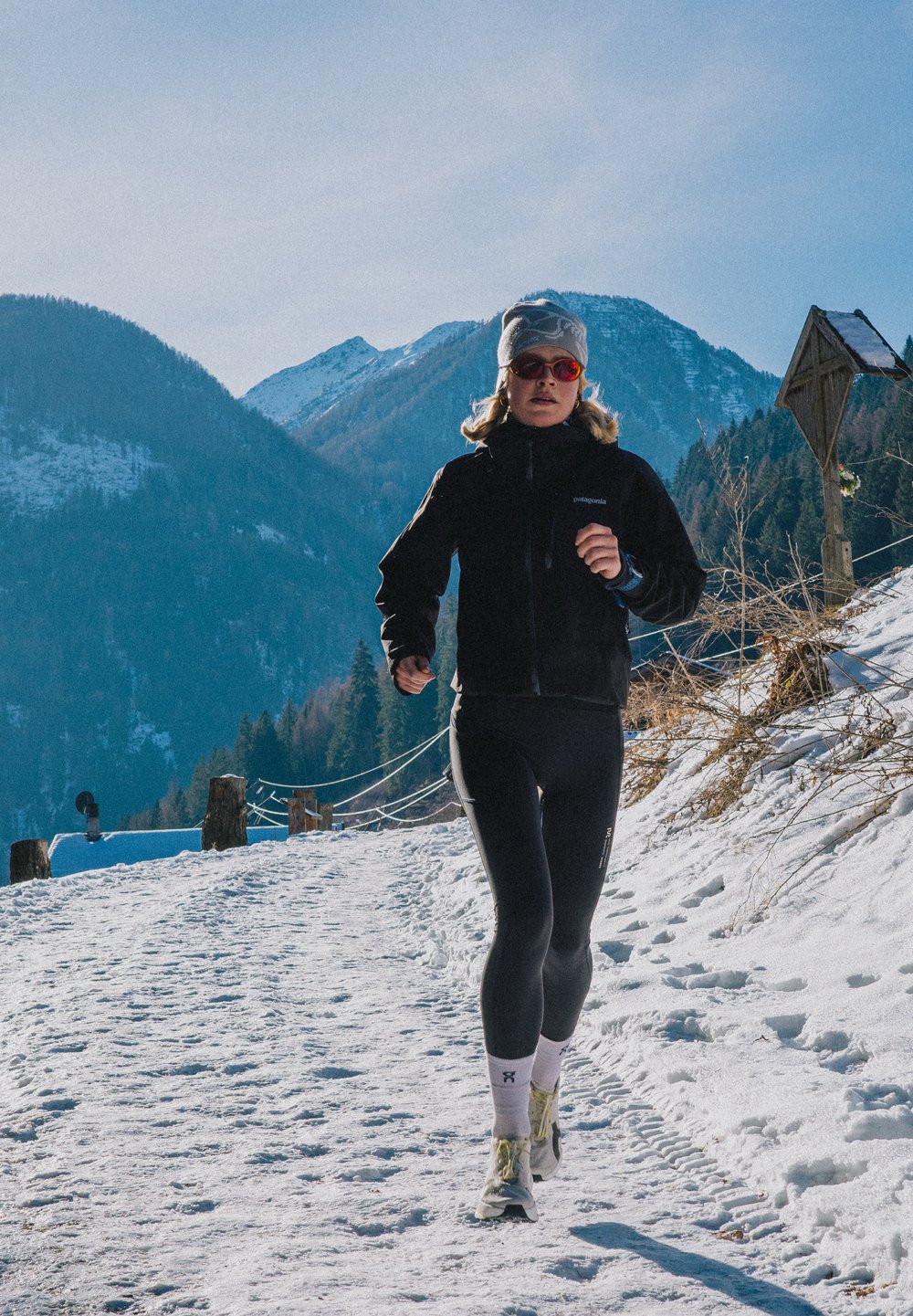 Veste noire isolante, leggings noirs, bonnet gris et lunettes de soleil rouges. Sentier enneigé et montagnes en arrière-plan.