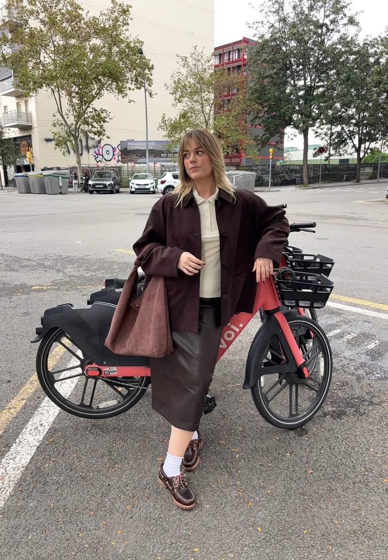 Bicycle électrique en noir et rouge avec un panier ; personne portant un manteau marron, une chemise beige, une jupe en cuir marron et des chaussures marron avec des chaussettes blanches.