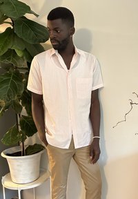 Camicia a maniche corte con bottoni di colore rosa chiaro con strisce bianche verticali, dotata di una tasca sul petto. Indossata con pantaloni aderenti beige.