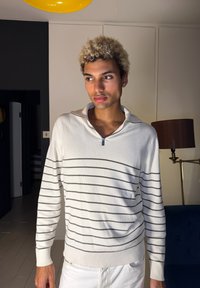 Jeune personne aux cheveux clairs et bouclés, vêtue d'un pull blanc rayé de fines bandes horizontales, debout dans un intérieur moderne.